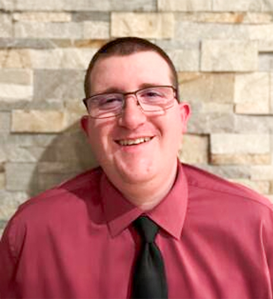 ID: White male wearing a red button down shirt, black tie and glasses smiling against a stone background.
