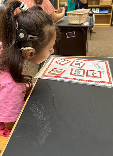 ID: An elementary student is working on a task that involves velcroed pictures. She has a ponytail and barretts holding her CI in place.
