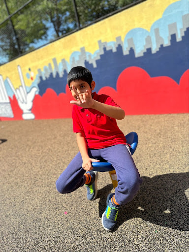 ID: An elementary student is sitting on a playground spinning chair. He is signing ILY.
