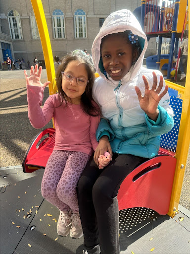 ID: Two young girls are holding hands and waving while sitting on a Merry Go Round seat together.
