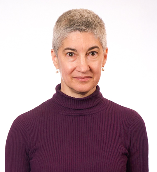 ID: A white woman with short salt & pepper hair wearing a purple turtleneck sweater and pearl drop earrings.