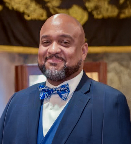 ID: A bald black man with a beard grinning and looking amused wearing a suit jacket and blue & white bowtie.