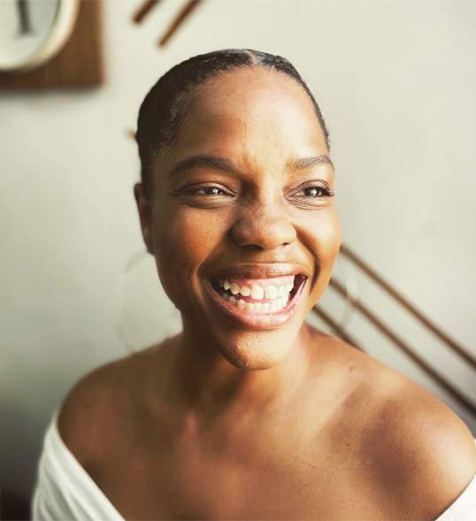 ID: A black woman wearing an off the shoulder top, smiling widely and looking to the side.
