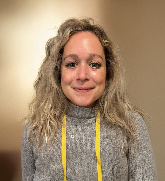 ID: A white woman with blonde wavy hair wearing a gray turtleneck sweater, silver necklace and yellow lanyard.