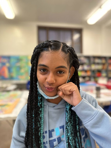 Young girl with long braided hair posing and smiling in a brightly lit classroom.